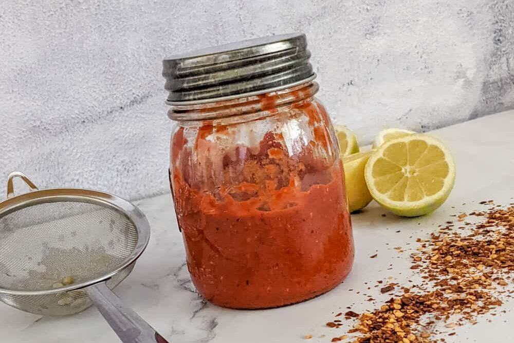 Homemade harissa sauce in a jar with lemons and red pepper flakes on a countertop.